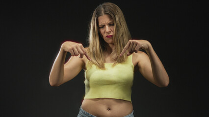 Woman points both index fingers down at navel wearing yellow tanktop in studio; disapproval warning.