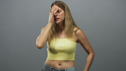 Fototapeta premium Woman in yellow crop top with hand pressed to forehead, other hand raised while turning head in studio; stress fatigue.