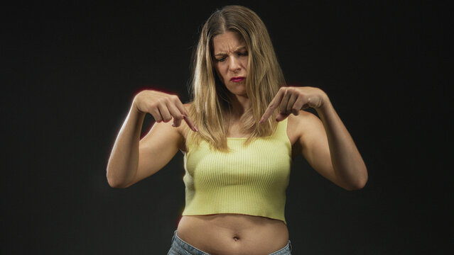 Woman points both index fingers down at navel wearing yellow tanktop in studio; disapproval warning.