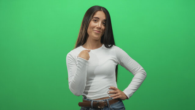 Woman with hand to chest and hand on hip in green studio backdrop, wearing white top and jeans, smiling and glancing upward; gratitude.