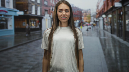 Fototapeta premium Teenage girl in white t shirt standing on a wet city street, grimacing and clenching fists with long hair down; frustration resilience.