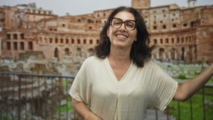 Fototapeta premium Middle age woman smiling, wearing glasses, hand on railing and arm outstretched, posing before an ancient ruin building; travel joy.