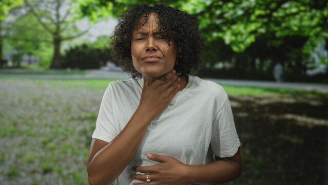 Woman wearing white t shirt clutching throat with one hand coughing in sunlit forest clearing; anxiety urgency health concern.