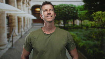 Obraz premium Man smiling and looking up beside a colonnade building with green park foliage and stone walkway, wearing olive tee; serenity health.