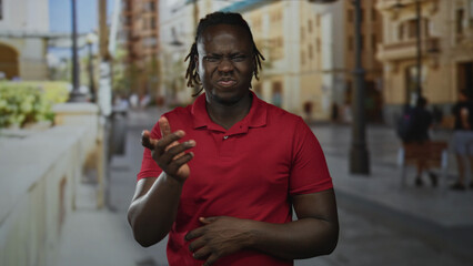 Fototapeta premium Young african american man in red shirt pinches nose with hand and grimaces on crowded street during daytime; disgust reaction.