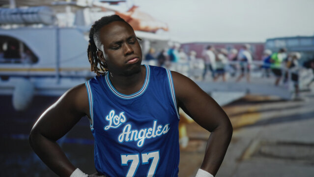 Man with hands on hips and bare forearms in blue basketball jersey on street pier near boats and dock; contemplation.