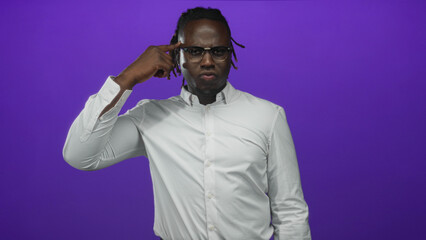 Fototapeta premium Young african american man pointing finger to temple while wearing glasses and white shirt in studio purple backdrop; thoughtful.