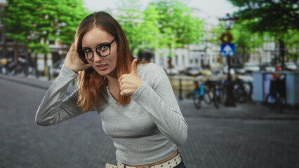 Fototapeta premium Young redhead woman with hand to ear giving thumbs up on a city street with bicycles and buildings visible; playful approval.