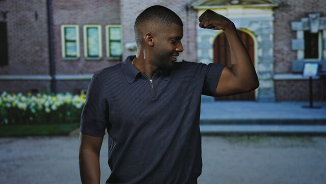Man flexes bicep with arm bent at elbow, smiling and looking at his raised arm beside a stone building entrance with steps and tulip flowerbed; confidence strength.