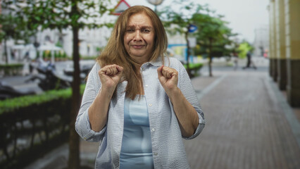 Fototapeta premium Woman with hands clenched near chest in a recoil gesture on a city street with sidewalk and parked scooters; unease caution.