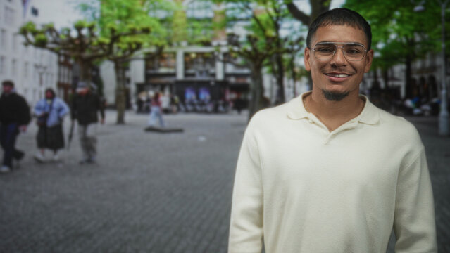 Man smiling with teeth visible, wearing glasses and a gold necklace, looking right while standing in a busy city street with trees and shops; casual contentment.
