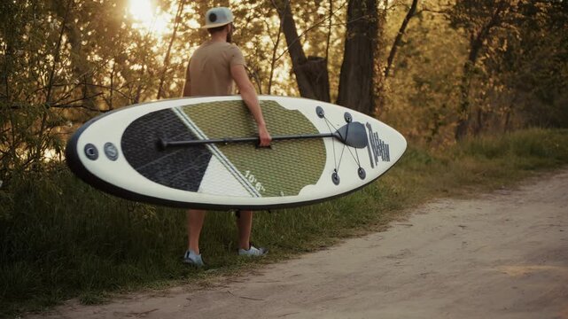 Man Carrying Stand Up Paddleboard Toward River At Sunset. Weekend Getaway 