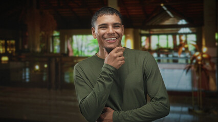 Naklejka premium Man touching mouth with his hand while smiling subtly in a building lobby; thoughtful quiet introspection.