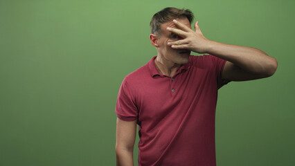 Fototapeta premium Man covering face with hand and peeking between fingers, wearing red polo against green screen in studio; embarrassment privacy vulnerability.