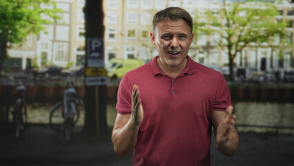Fototapeta premium Man clapping hands beside parked bicycles on a canal street in front of brick townhouses, red polo visible; engaging presentation.