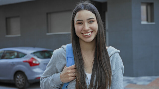 Young woman student with blue backpack holds strap and smiles facing camera on a city street; youthful optimism.