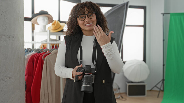 Woman holding camera and gesturing with open hand in studio among lights and clothing rack; confidence approachability.