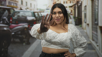 Fototapeta premium Woman showing four fingers with outstretched hand and slight smile, wearing white embroidered crop top and nose ring while standing on a narrow city street; playful confidence.