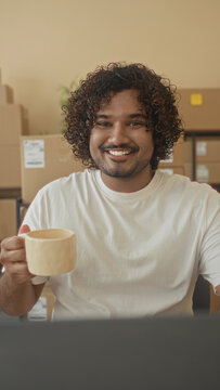 Man holding coffee cup, hand raised to sip in building office with cardboard boxes and laptop; small business optimism.