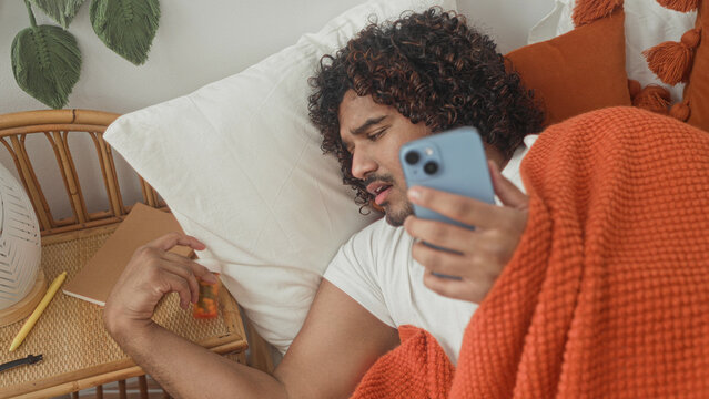 Man in bed holding smartphone, hand on neck and reaching for pill bottle on bedside table, covered by orange blanket, studio; sick.