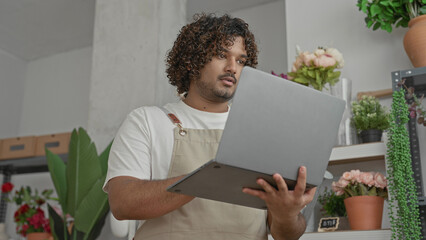 Man florist in apron holds laptop and stylus and points at screen in building; focus inventory...