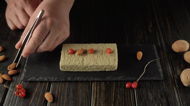 Hands carefully position almonds and red berries on a textured block halva of food placed on a dark slate plate, surrounded by scattered almonds and berries on a wooden surface