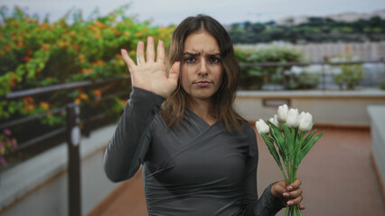 Obraz premium Woman holding white tulips on building terrace raises open hand palm toward camera in a clear stop gesture, frowning slightly; firm refusal.