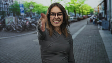 Fototapeta premium Young hispanic woman extends hand for handshake on city street, smiling and wearing glasses and grey top; friendly greeting.