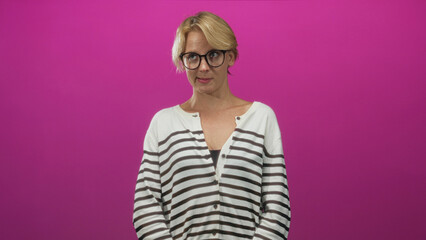 Fototapeta premium Woman with blond hair and round glasses looking down with hands clasped wearing a striped sweater in studio; quiet contemplation.