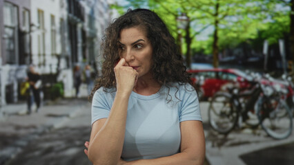 Fototapeta premium Woman standing and touching her nose with her finger under green trees beside parked bicycles on a city street; doubt.