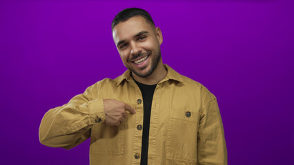 Obraz premium Man wearing tan jacket smiles and points finger to shirt while giving thumbs up in purple studio; confidence.
