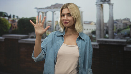 Fototapeta premium Woman holds up hand showing palm and fingers while slightly smiling in front of stone columns on a building terrace, denim shirt visible; friendly greeting.
