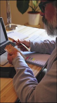 Serious senior caucasian man with a beard and eyeglasses managing home expenses, counting euro banknotes, and using a laptop computer while sitting at his wooden desk in a modern interior