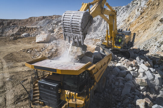 Aerial view of a large yellow excavator with a worker in the cab loading heavy rocks into a massive dumper truck at an open-pit mine Santa Lucia de Gordon, Castile and Leon, Spain.