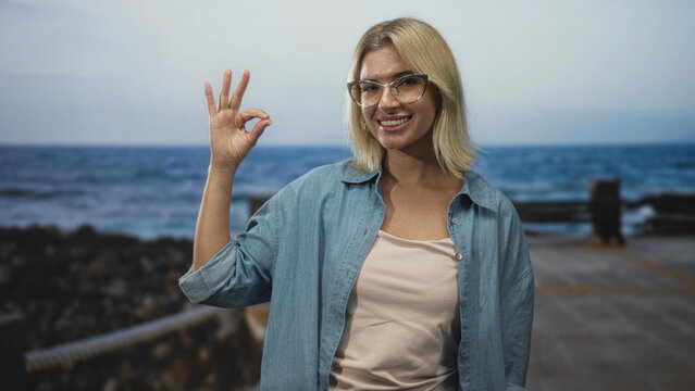 Woman smiling and holding hand in ok sign in a studio with pier backdrop and denim shirt visible; confidence approval.