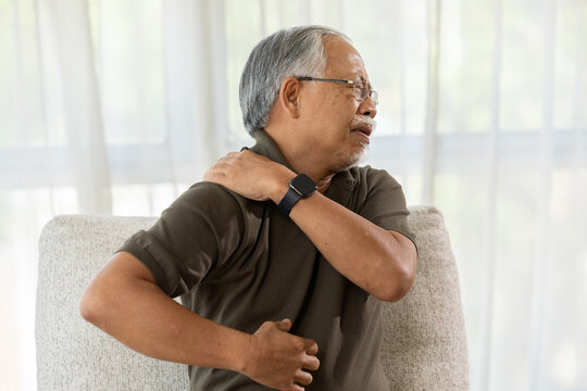 Senior man experiences shoulder pain massaging his aching neck feeling uncomfortable while sitting on sofa at home living room showing painful expression
