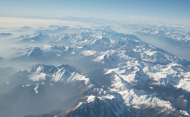 Naklejka premium Airplane window view of snowy Alpine mountains above clouds during flight over Europe.