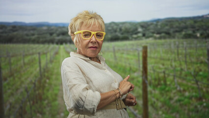 Fototapeta premium Woman points finger and checks wrist in a vineyard wearing yellow glasses and linen dress; thoughtful reflection.