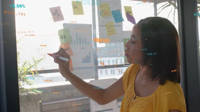 Woman reaching and writing on glass circling biz data while noting insight with overlays appearing