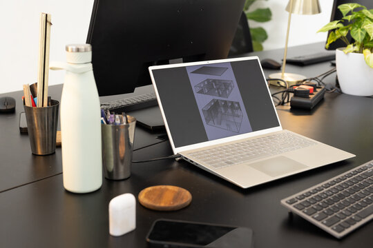 Laptop displaying CAD drawings on dark wood desk, showing keyboard, monitor, bottle, plant