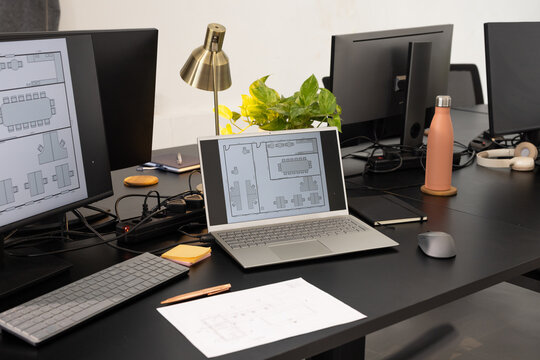 Silver laptop displaying floor plan on black desk in office with monitors, coral water bottle