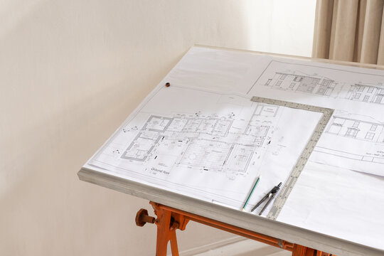 Drafting table holding plans, T-square, compass, pencil on alum edge with orange sawhorse in studio