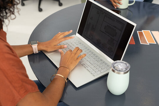 Coworkers, woman wearing rust top, pink watch working on laptop in studio with layout and swatches