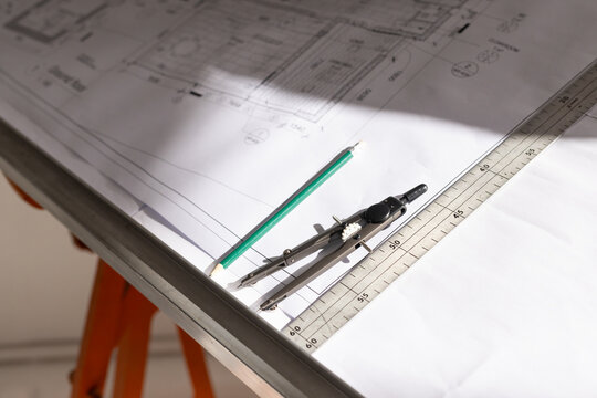 Metal ruler, compasses and green pencil are resting on blueprint on drafting table with shadow