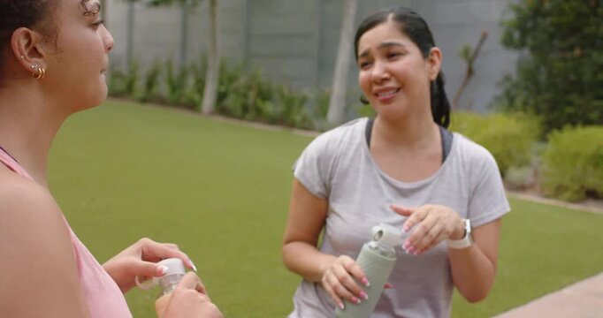 Diverse female friends in sportswear standing in yard left sipping bottle urging right showing cap