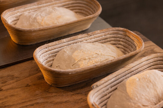 Oval woven bannetons holding three flour-dusted loaves of proofing dough on wooden countertop
