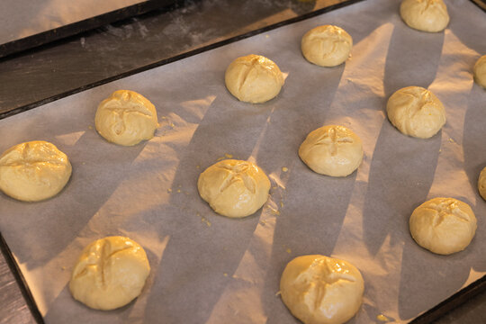 Metal tray lined with parchment paper holding scored dough buns shiny from eggwash on countertop