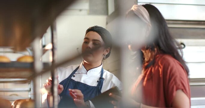 Diverse female coworkers checking rack in bakery, manager pointing tablet, chef in coat adjusting