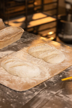 Two scored wheat loaves proofing on floured wooden peel at bakery prep table with yellow scraper