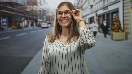Fototapeta premium Woman wearing striped shirt adjusts her round glasses with a gentle smile while standing on a bustling city street; confidence.
